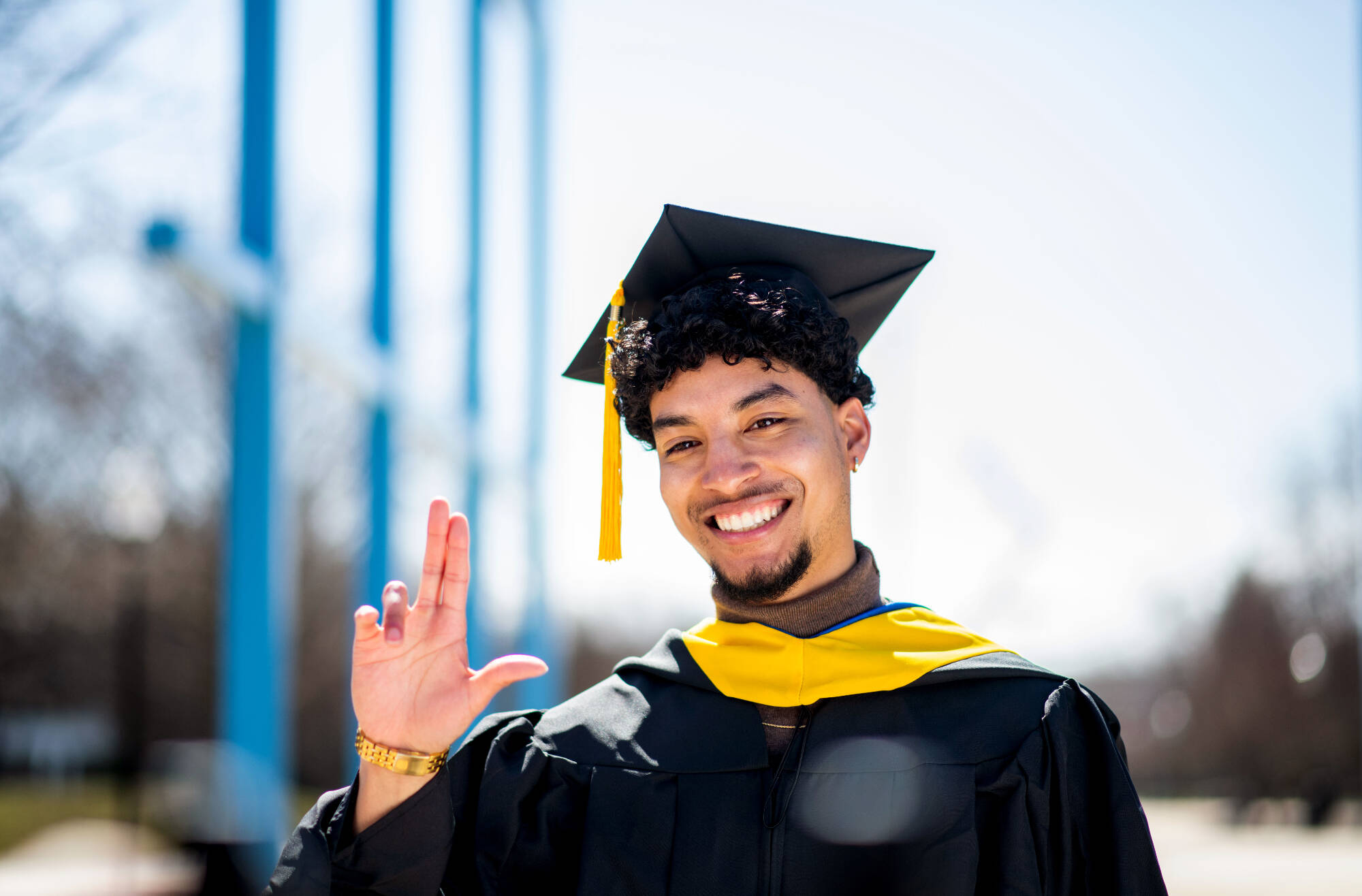 A GVSU Graduate doing the Anchor Up hand signal in front of the Transformational Link. He is wearing his cap and gown.